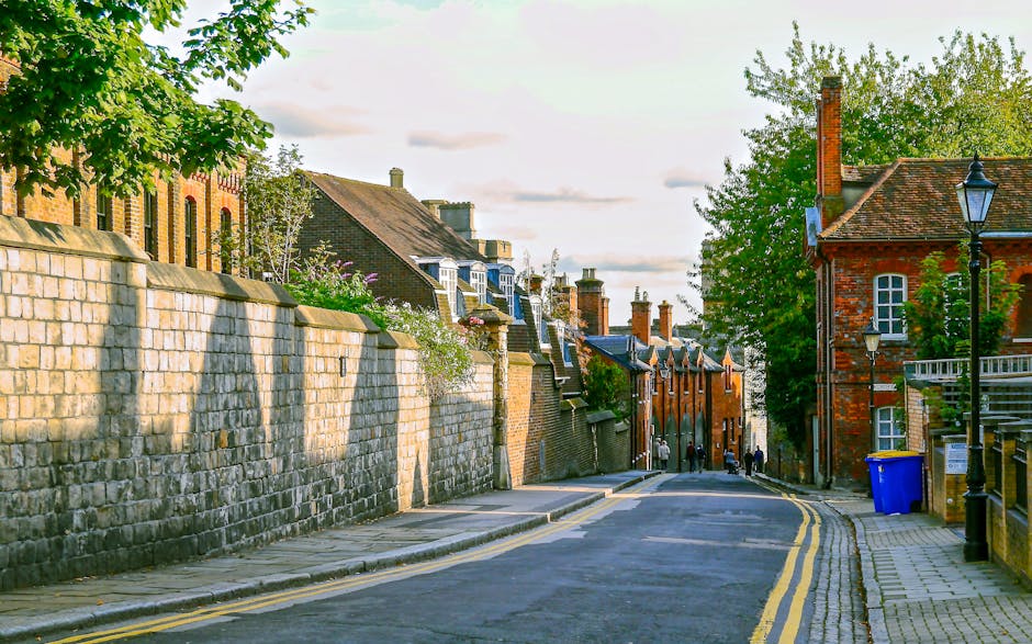 A narrow alleyway between two brick buildings featuring textured red brick walls with small, rectangular windows and black drainage pipes running vertically along the facades. The concrete pavement st